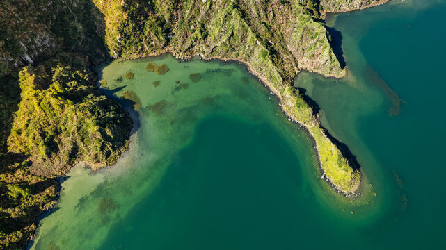 Aerial view of dramatic emerald waters embracing a rugged, verdant coastline with a slender island, Ponta Delgada, Azores, Portugal.