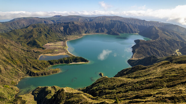 Aerial view of the stunning emerald-hued caldera lake nestled amidst rugged, verdant slopes under a bright azure sky, Ponta Delgada, Azores, Portugal.