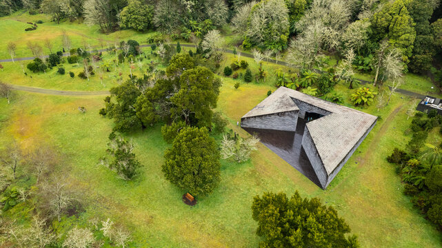 Aerial view of a striking dark angular building with a central void, nestled amidst vibrant green lawns and diverse trees, Ponta Delgada, Azores, Portugal.