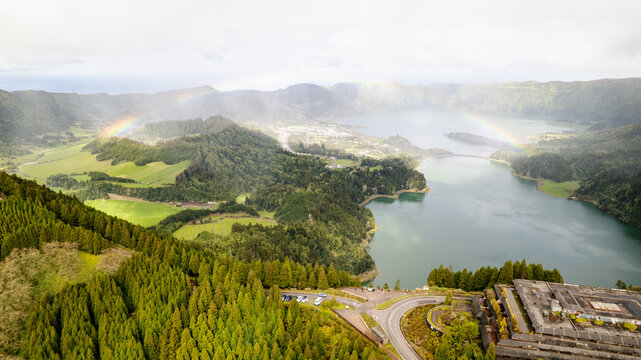 Aerial view of the twin lakes reflecting the sky, separated by a narrow strip of land, with a rainbow arching above the lush green landscape, Ponta Delgada, Azores, Portugal.