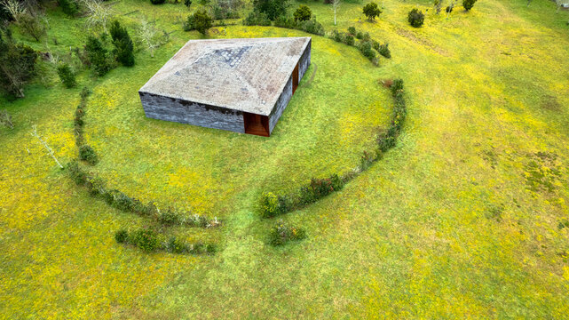 Aerial view of a lone, dark-roofed structure amidst bright green and yellow fields, encircled by a low stone barrier, Ponta Delgada, Azores, Portugal.