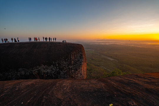 A peaceful morning at Three Whale Rock, where visitors gather on rugged cliffs to witness the first light of day. Misty valleys, dense forests, and glowing skies create a dreamlike natural panorama.