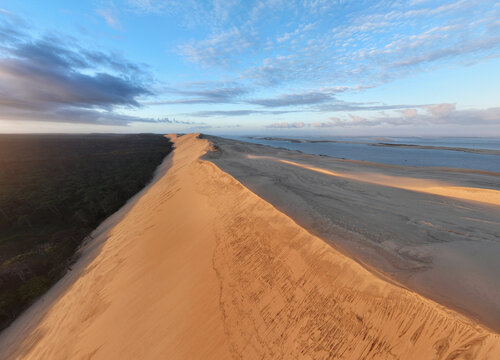 Aerial view of the colossal golden Dune du Pilat dramatically meeting the dark, dense forest and shimmering bay under a vast, cloud-streaked sky, La Teste de Buch, Nouvelle-Aquitaine, France.