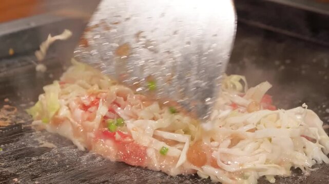 High-angle shot of a chef or diner preparing traditional Japanese savory pancakes (Okonomiyaki) or pan-fried batter (Monjayaki) on a hot teppan griddle. The footage captures the steam rising from a mi