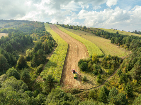 Aerial view of striped green and golden fields carved into rolling hills, where a lone tractor harvests crops under a dramatic sky, Tuzla, Bosnia and Herzegovina.