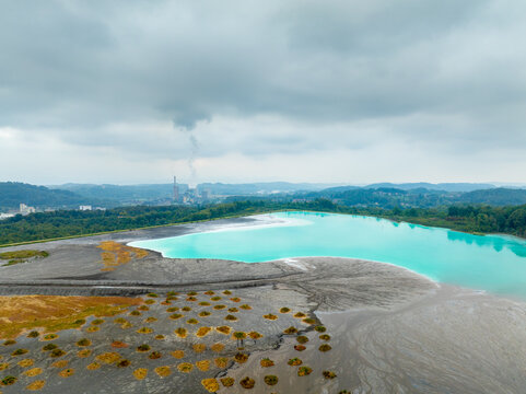 Aerial view of a vibrant turquoise industrial pond starkly contrasting with barren grey land dotted with orange patches, under a heavy sky with distant smoking chimneys, Tuzla, Bosnia and Herzegovina.
