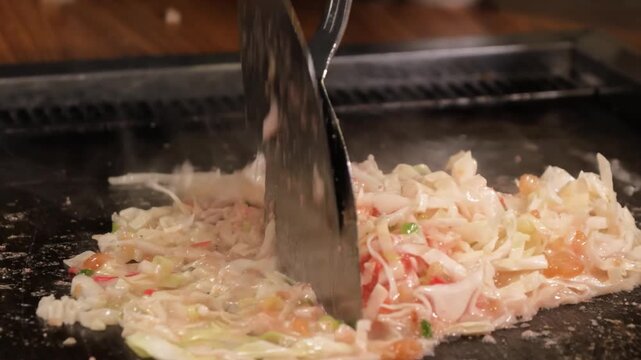 close-up of a chef or diner preparing Monjayaki, a traditional Tokyo soul food, on a hot teppan iron plate. The footage captures the steam and sizzle of the thin, runny batter mixing with finely chopp