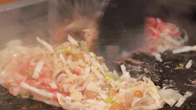 Close-up footage of traditional Japanese Monjayaki being prepared on a hot iron teppan grill. The scene shows the characteristic runny batter mixing with finely shredded cabbage and red ingredients, t