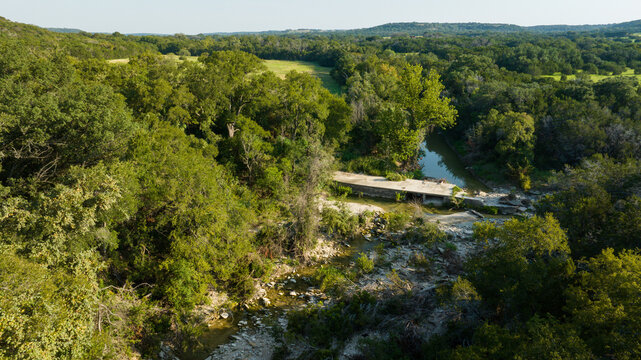 Aerial view of a weathered concrete bridge spanning a tranquil river, cutting through a vibrant tapestry of sun-drenched green foliage, Freeport, Texas, United States.