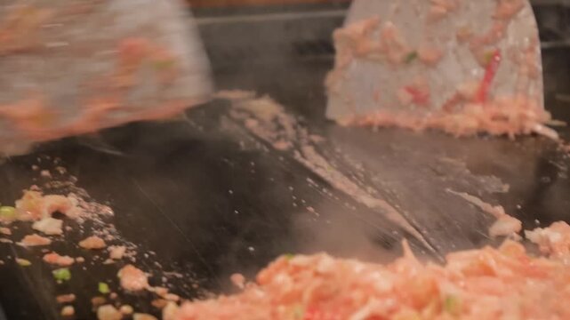 High-angle close-up shot of a skilled chef or diner preparing Monjayaki, a savory pan-fried batter dish popular in Tokyo's downtown districts. The footage captures the rhythmic chopping of fresh ingre