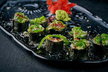 Sushi maki rolls topped with seaweed salad (chuka), sesame seeds, and balsamic glaze, served with ginger and wasabi on a dark plate © ismishko