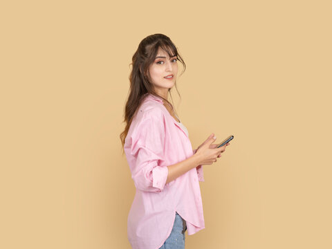 Young woman holding smartphone and looking over shoulder with neutral curious expression. Isolated on beige background copy space, suitable for mobile usage, communication and lifestyle concepts.