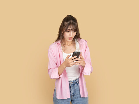 Worried young woman looking at smartphone with anxious expression while reading message. Isolated on beige background with copy space, ideal for problem, bad news or stress concept.