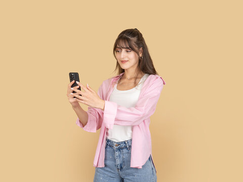 Smiling young woman holding smartphone and taking photo or selfie, looking at screen with pleasant expression. Isolated on beige background, copy space for social media and digital lifestyle concepts.