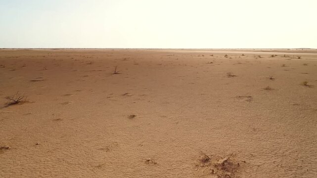 Aerial slow tracking forward shot of cracked desert plain with sparse shrubs and parched soil under bright midday sky across flat remote landscape