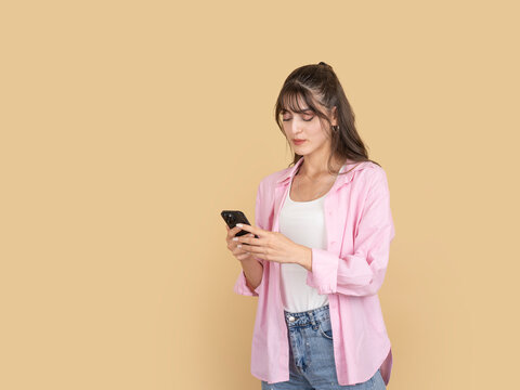 Young woman in pink shirt using smartphone with calm focused expression, typing message on beige background, copy space. Ideal for mobile communication, social media and digital lifestyle concepts.