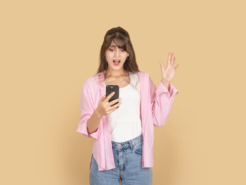 Shocked young woman looking at smartphone with raised hand and surprised expression. Isolated on beige background with copy space, ideal for unexpected message, problem or breaking news concept.