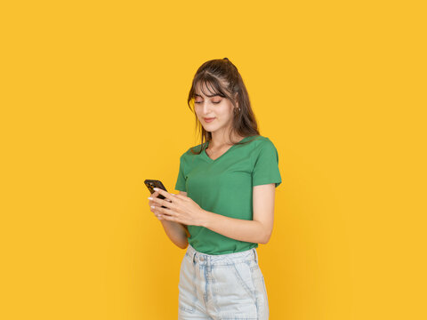 Young woman using smartphone with focused expression, typing message while standing over yellow background with copy space. Ideal for mobile communication, social media, digital lifestyle concepts.