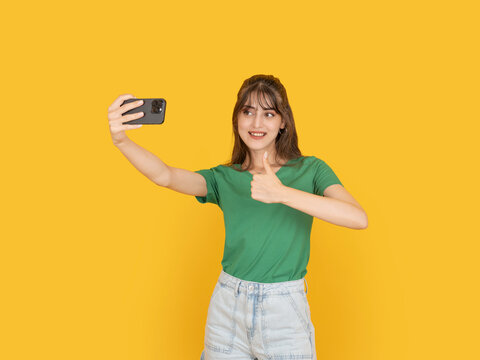 Young woman taking selfie with smartphone while showing thumbs up gesture. Isolated on bright yellow background with copy space, ideal for social media, approval or lifestyle concept.