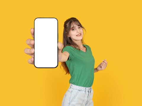 Smiling young woman holding smartphone with blank screen toward camera in dynamic pose. Isolated on bright yellow background with copy space, ideal for mobile app promotion or advertising mock up.