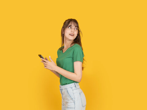 Young woman holding smartphone and looking aside with curious expression. Isolated on bright yellow background with copy space, ideal for lifestyle, communication or mobile usage concept.