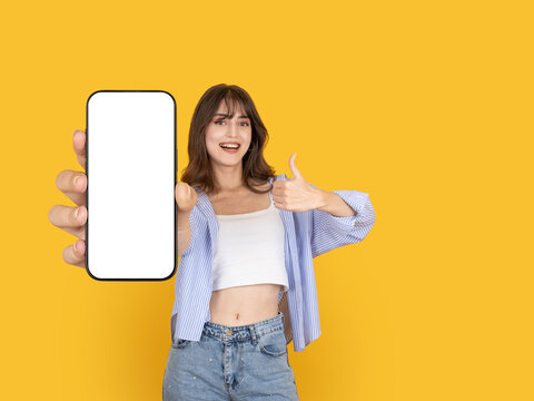 Cheerful young woman holding smartphone with blank screen and showing thumbs up gesture. Isolated on bright yellow background with copy space, ideal for mobile app promotion or advertising mock up.