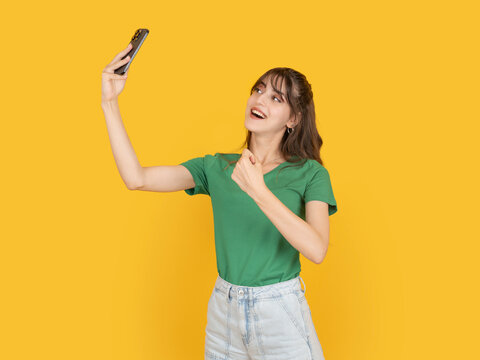 Excited young woman holding smartphone up for selfie and clenching fist in celebration, reacting to success or good news. Isolated on yellow background with copy space for advertising use.
