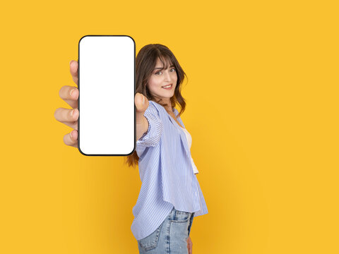 Smiling young woman holding smartphone with blank screen toward camera in close perspective. Isolated on bright yellow background with copy space, ideal for mobile app or advertising mock up concept.