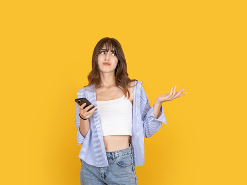 Confused young woman holding smartphone and raising hand with questioning expression, reacting to unclear message or problem. Isolated on yellow background with copy space for advertising use.