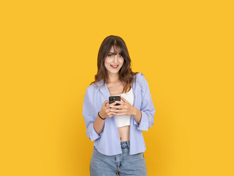 Smiling young woman holding smartphone with both hands and looking at camera with confident friendly expression concept idea image. Isolated on yellow background with copy space for advertising use.