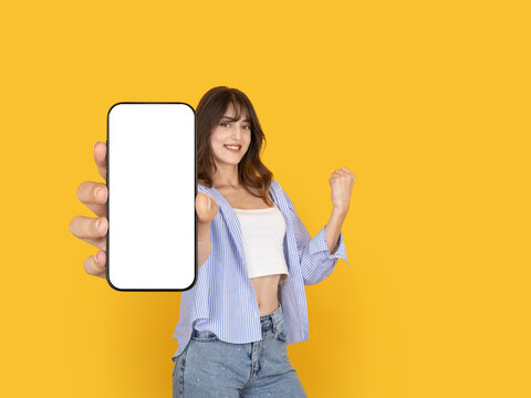 Confident young woman showing smartphone mock up with blank screen to camera and celebrating success with raised fist concept. Isolated on bright yellow background with copy space for advertising use.