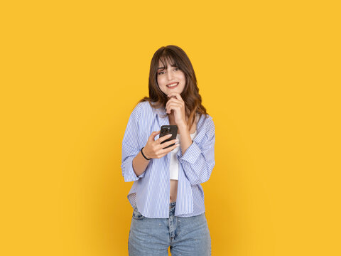 Thoughtful smiling young woman holding smartphone and touching chin with curious expression, considering message or idea concept. Isolated on yellow background with copy space for advertising use.