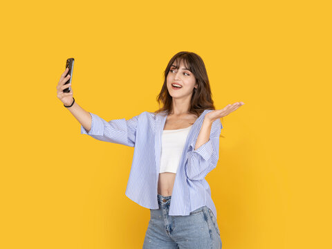 Smiling young woman taking selfie with smartphone while gesturing open hand in presentation pose, expressing excitement and engagement. Isolated on yellow background with copy space, advertising use.