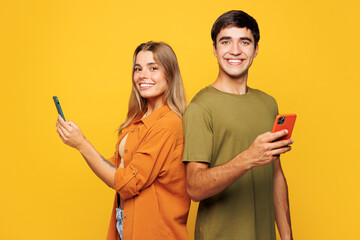 Naklejka premium Side view smiling young couple two friends family man woman wear casual clothes hold in hand use mobile cell phone together stand back to back isolated on pastel plain light yellow background studio.