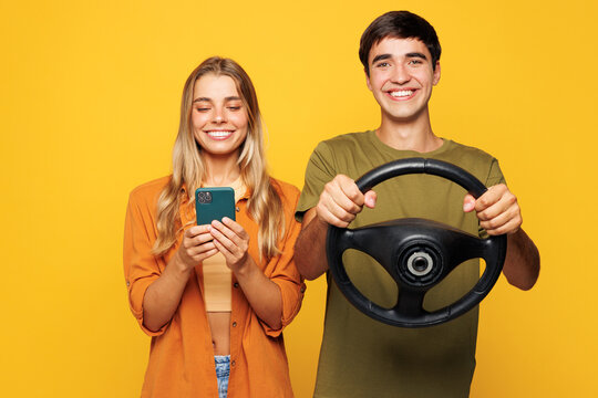 Young couple two friends family man woman wears shirt casual clothes together hold steering wheel driving car use mobile cell phone isolated on pastel plain light yellow background. Lifestyle concept.