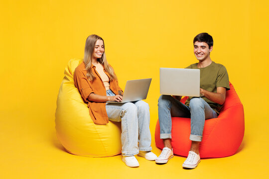 Full body smart young couple two friends family IT man woman wear pink casual clothes together sit in bag chair hold work on laptop pc computer isolated on plain yellow background. Lifestyle concept.