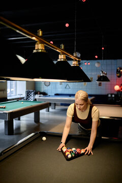Vertical shot of Caucasian young woman arranging billiard balls on pool table in recreation venue, preparing game start. Suitable for leisure marketing, nightlife promotion, social activity ads
