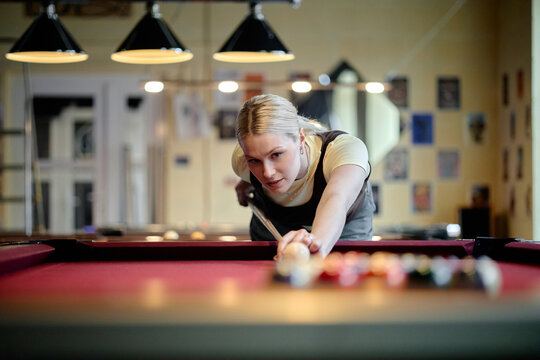 Young adult Caucasian woman aiming pool cue and lining up shot on billiards table, showing focus and leisure activity. Useful for recreation marketing, nightlife promotion, hobby content
