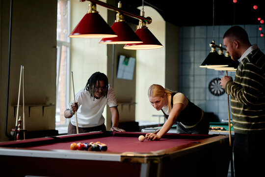 Young adult friends playing billiards in social club, aiming shot and watching game together. Useful for leisure marketing, team bonding, nightlife promotion, medium shot
