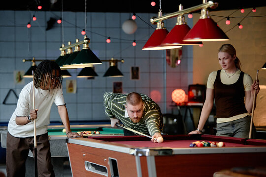 Young adult friends playing billiards in entertainment venue, taking shot and socializing during leisure time. Useful for youth lifestyle, recreation, teamwork, nightlife marketing
