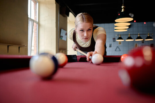 Portrait of Caucasian young woman aiming cue ball on billiards table, showing focus, precision, leisure competition. Useful for recreation marketing, youth lifestyle campaigns, close up
