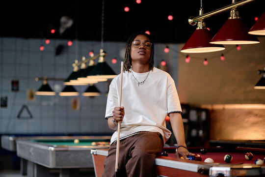 Portrait of young adult Black man holding pool cue while sitting on billiards table, representing leisure venue culture and social entertainment. Useful for nightlife marketing and youth lifestyle cam