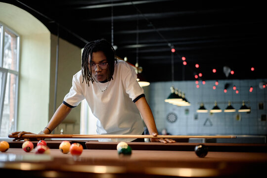 Young Black man aiming pool shot in recreation venue, focusing on leisure activity and social entertainment. Useful for youth lifestyle, hobby marketing, nightlife promotion campaigns
