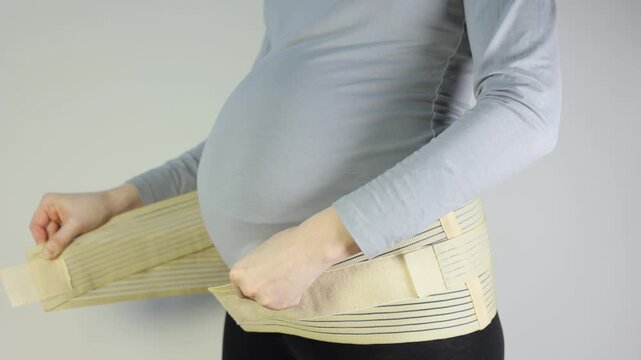 Pregnant woman adjusting a supportive maternity belt over her grey shirt to alleviate back pain and provide abdominal stability during pregnancy.