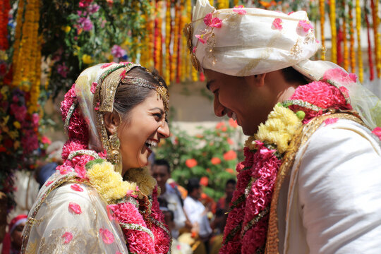 Professional scene of indian bride and groom laughing together while exchanging flower garlands during varmala ceremony, colorful flowers and festive decorations, bright natural daylight for social