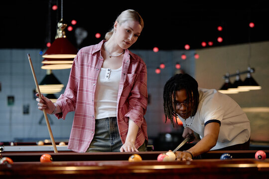 Young adult Caucasian woman and Black man playing pool in billiards club, aiming shot and socializing. Usable for nightlife marketing, leisure campaigns, dating app or friendship promotion

