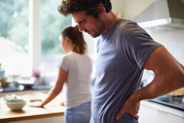 Fototapeta premium Professional scene of man holding lower back in pain while standing in kitchen, concerned partner nearby offering help, bright home interior, natural daylight from window, for web design