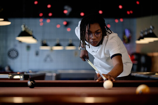 African American teenager playing billiards, aiming cue ball in pool hall and focusing on leisure competition. Usable for youth lifestyle, recreation marketing, social campaigns
