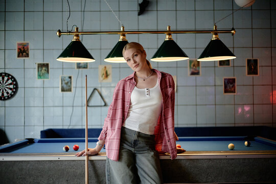 Portrait of Caucasian young adult woman posing with billiard cue beside pool table, suggesting leisure venue marketing. Useful for entertainment promotion, nightlife ads, social campaigns
