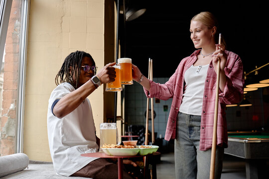 Young adult Black man and Caucasian woman clinking beer mugs while playing pool in bar, socializing after work. Ideal for leisure marketing, friendship, nightlife promotion

